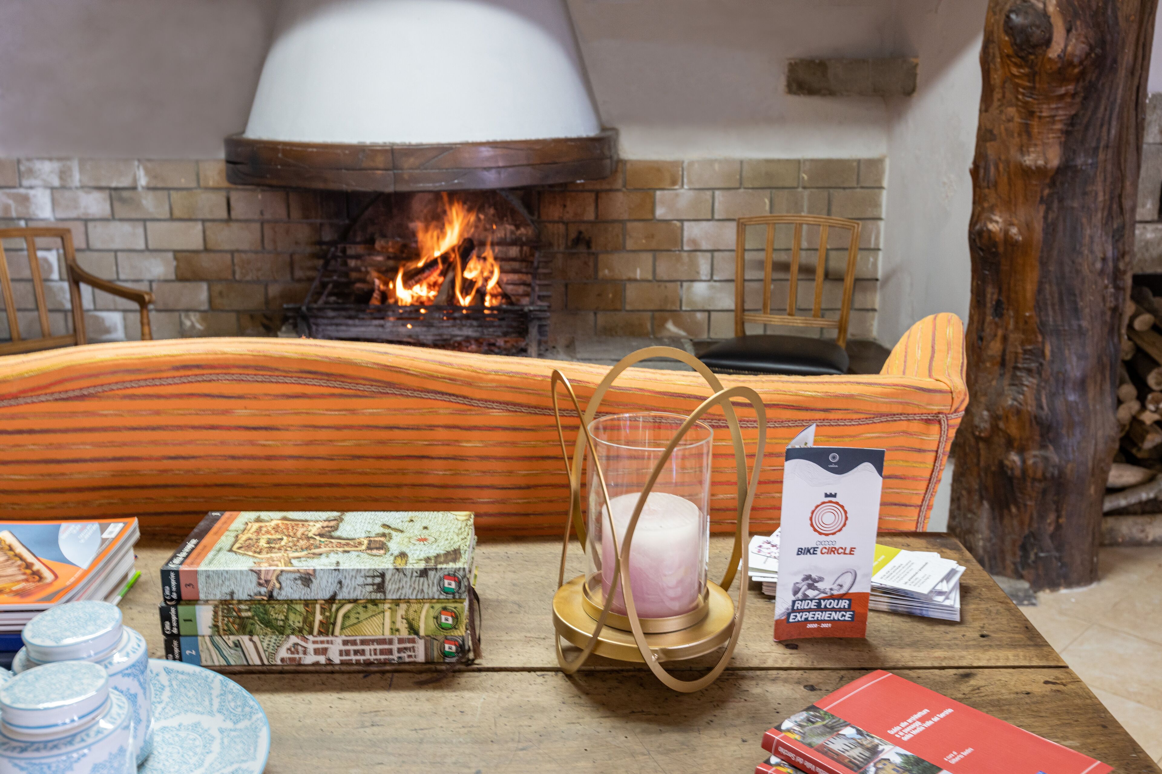 Une pièce chaleureuse avec une cheminée en briques, un banc en bois et une table sur laquelle on trouve des livres, une bougie et un sac de collations.