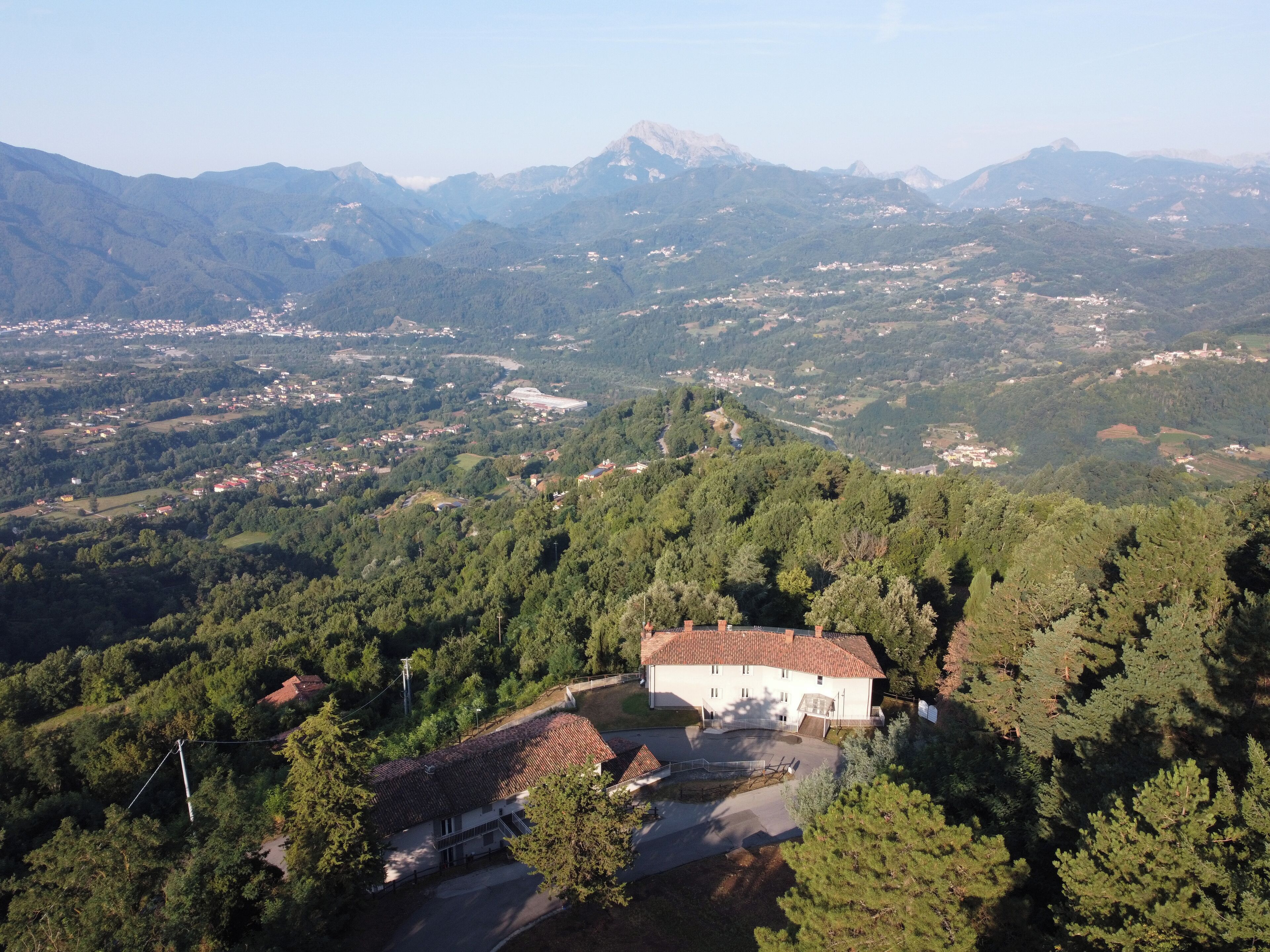 A rural house with a tiled roof surrounded by dense forest, with mountains in the background.