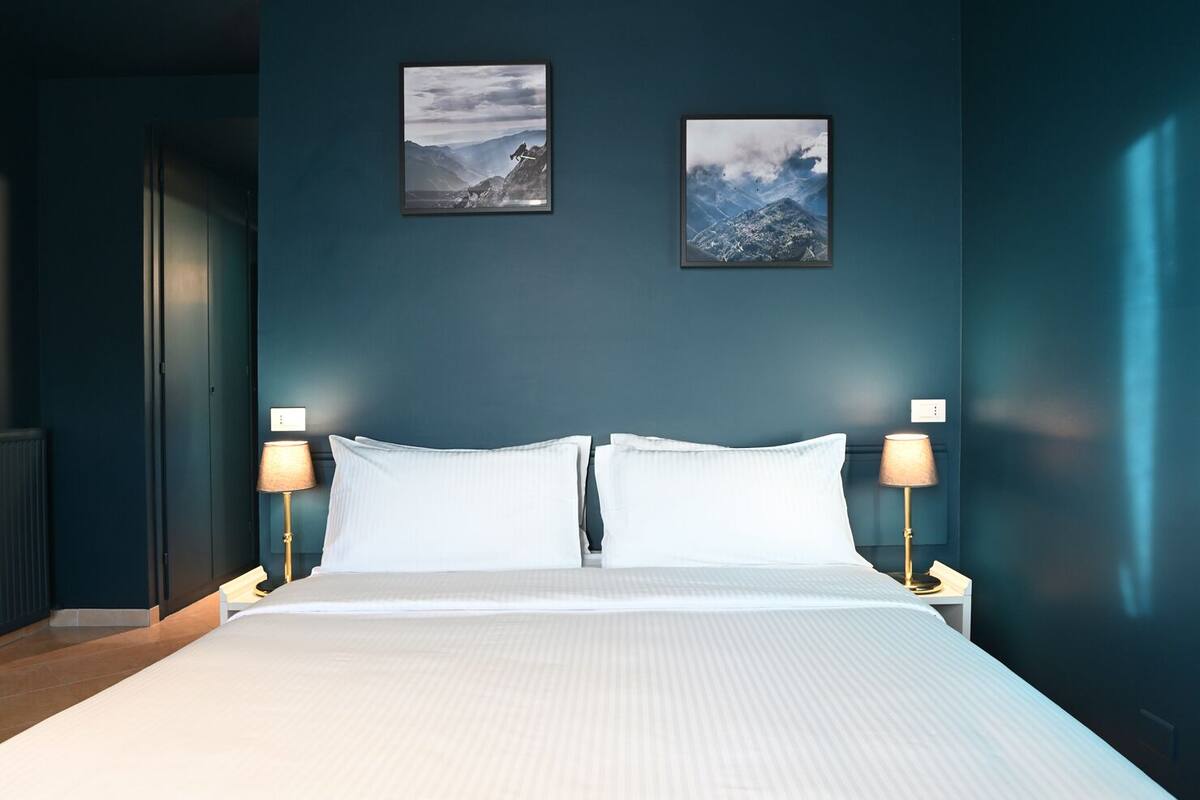 A neatly made bed with white linens in a room with dark blue walls. There are two framed pictures on the wall above the bed.
