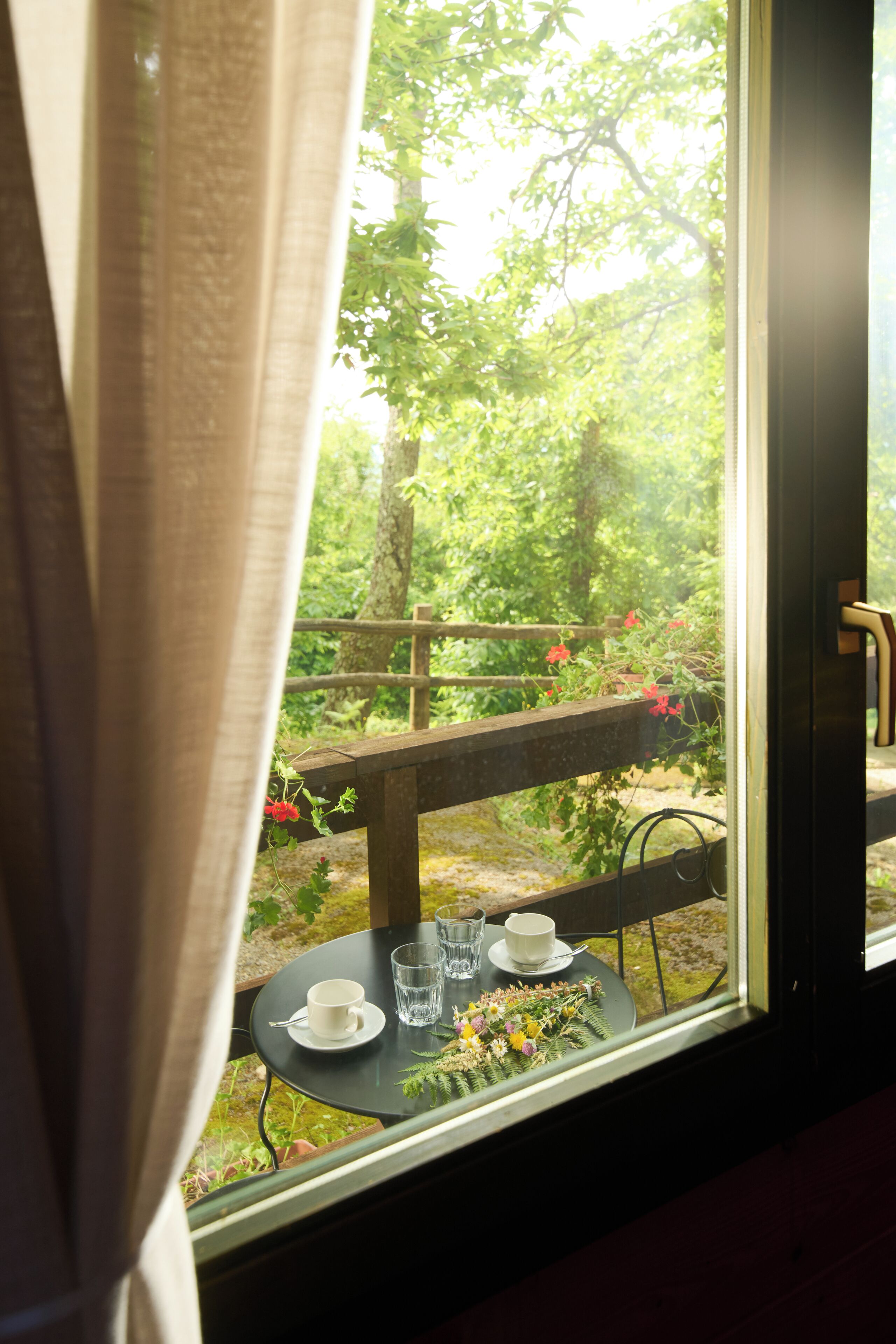 Una ventana con vista a una terraza de madera, una mesa con una taza y un vaso, y una cortina entreabierta.