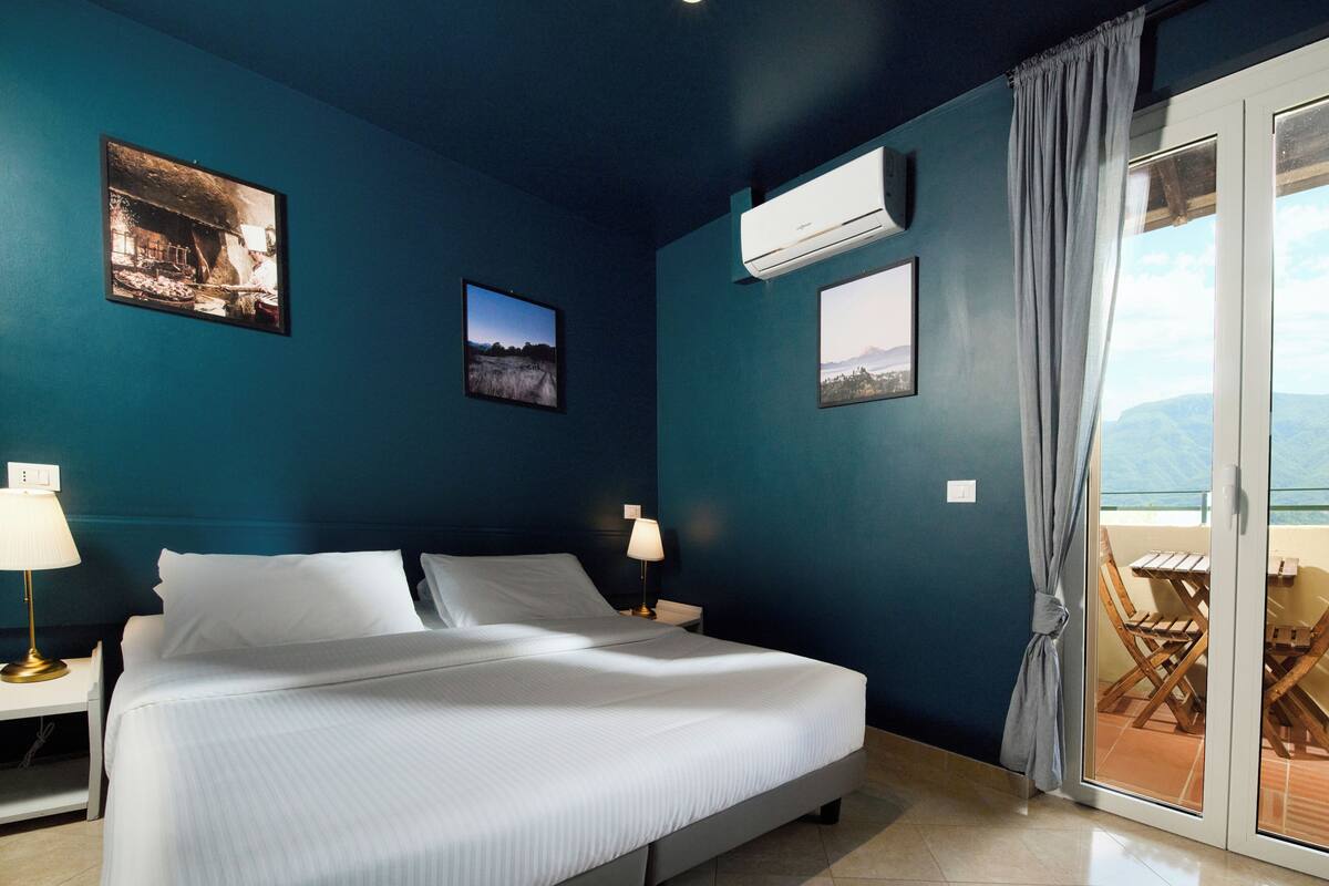 A bedroom with a bed, two nightstands, a window with a view, and an air conditioning unit.