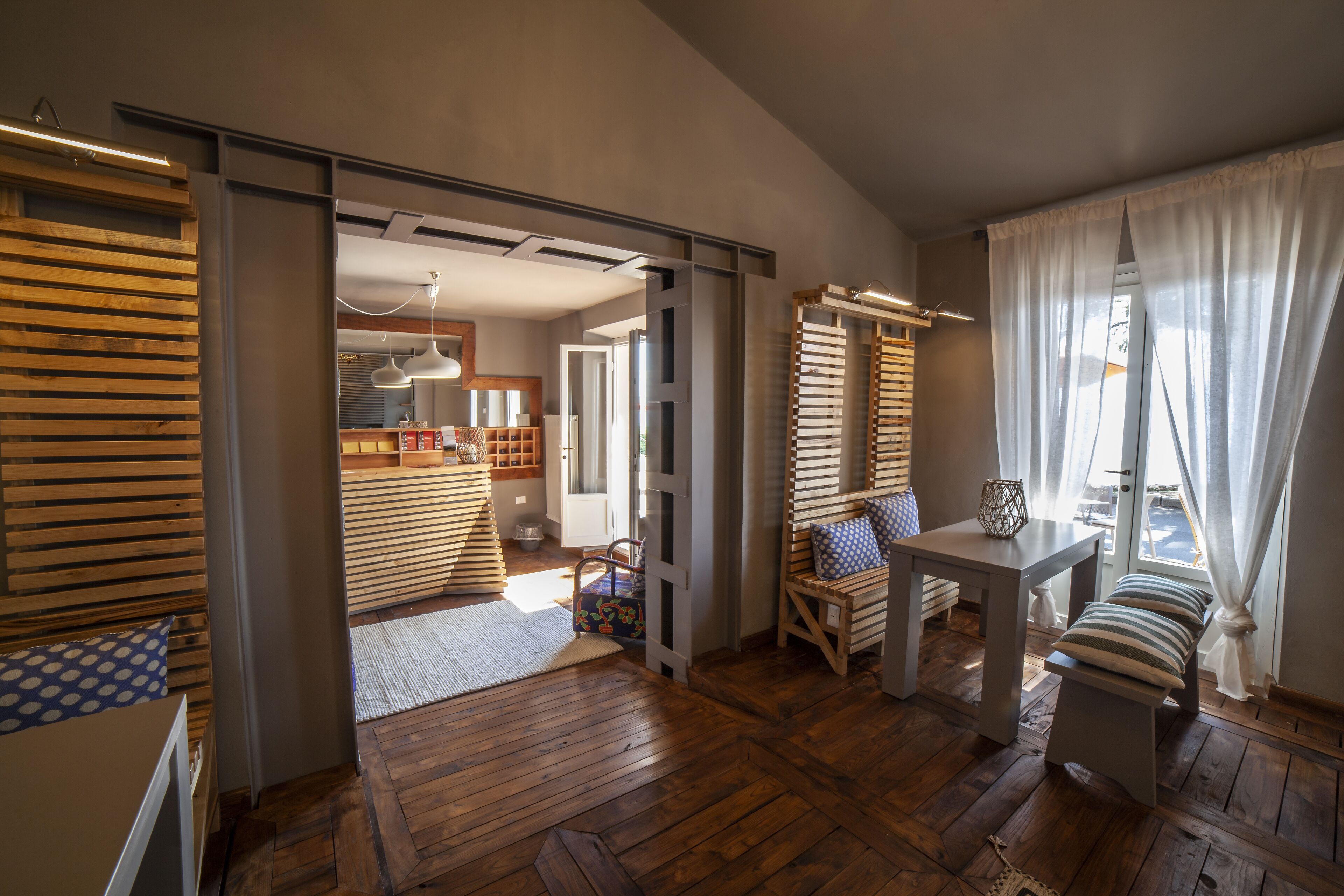 A spacious room with wooden flooring, a bench, a table, and a view through a window with sheer curtains.