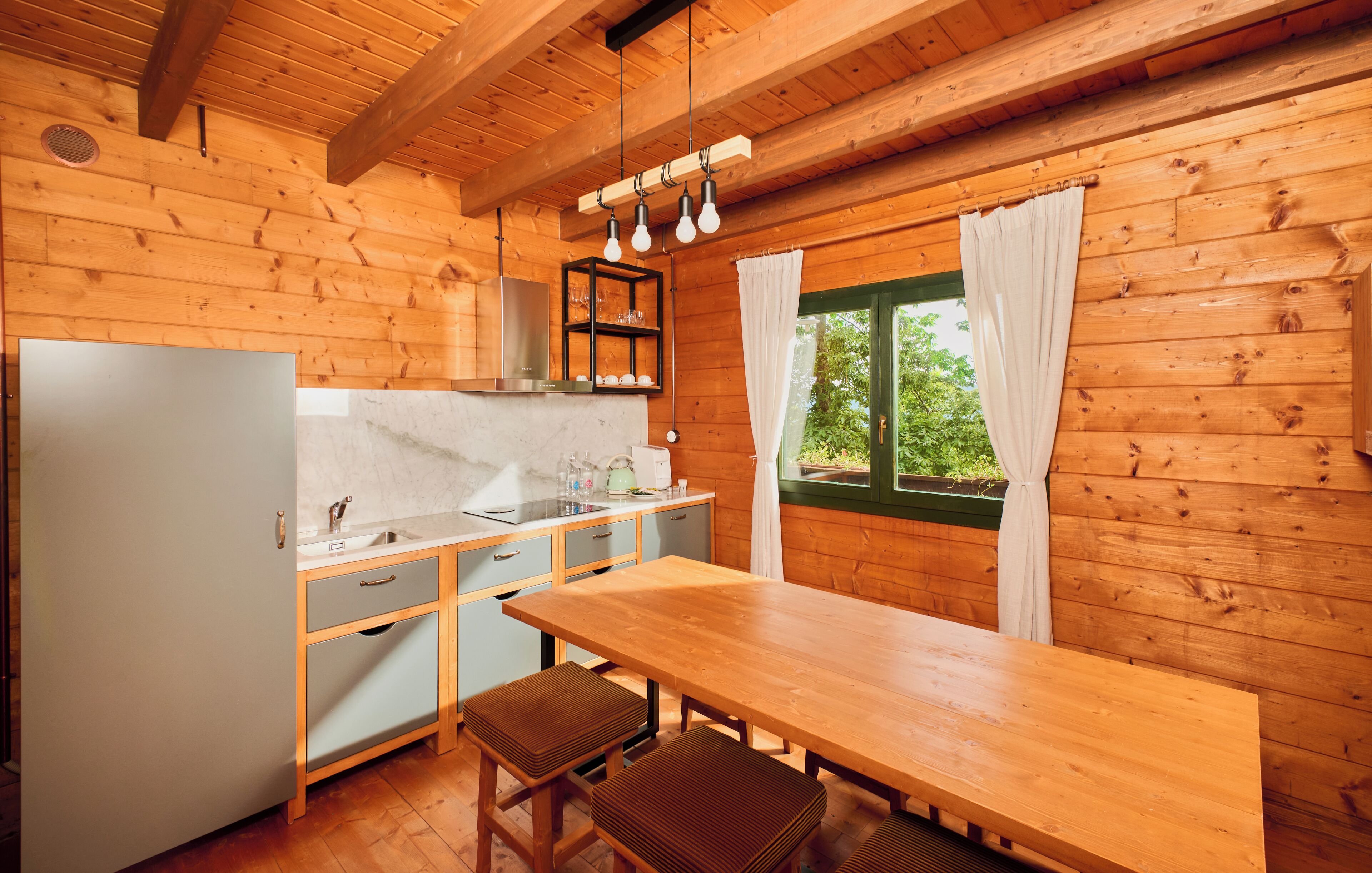 Eine Holzhütte mit einer Küche, einem Holztisch, zwei Barhocker, einem Spülbecken und einem Fenster mit Vorhängen.