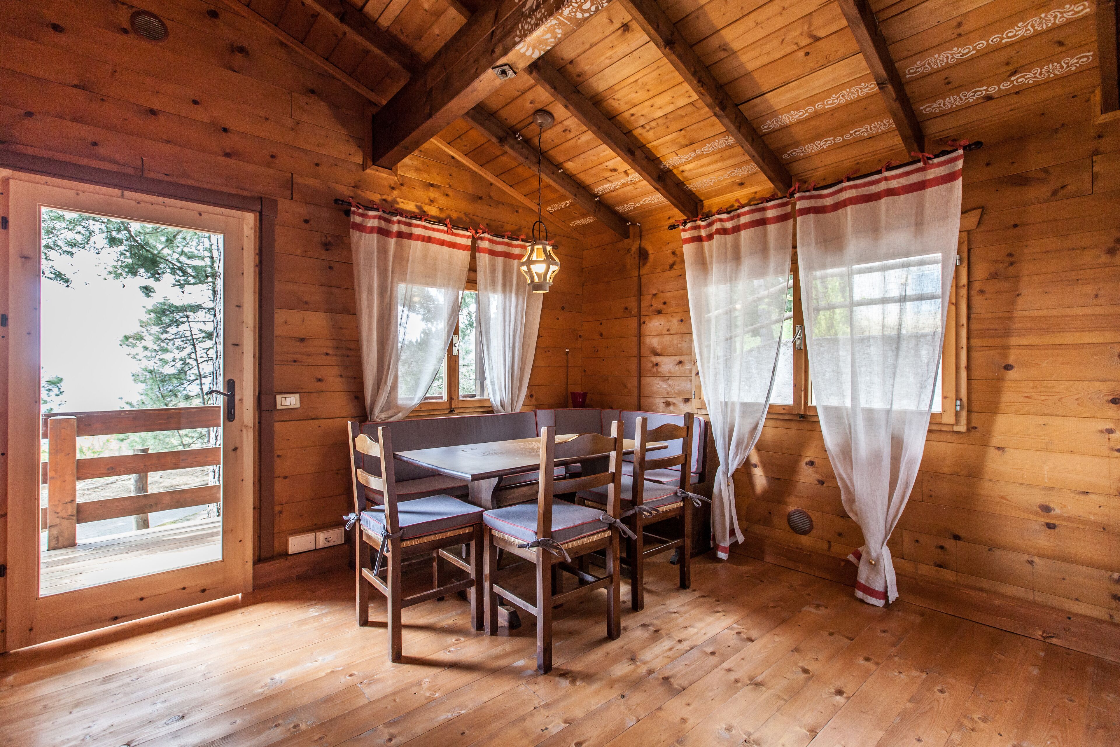 A wooden cabin with a dining table and chairs, a wooden ceiling with beams, and a window with curtains.