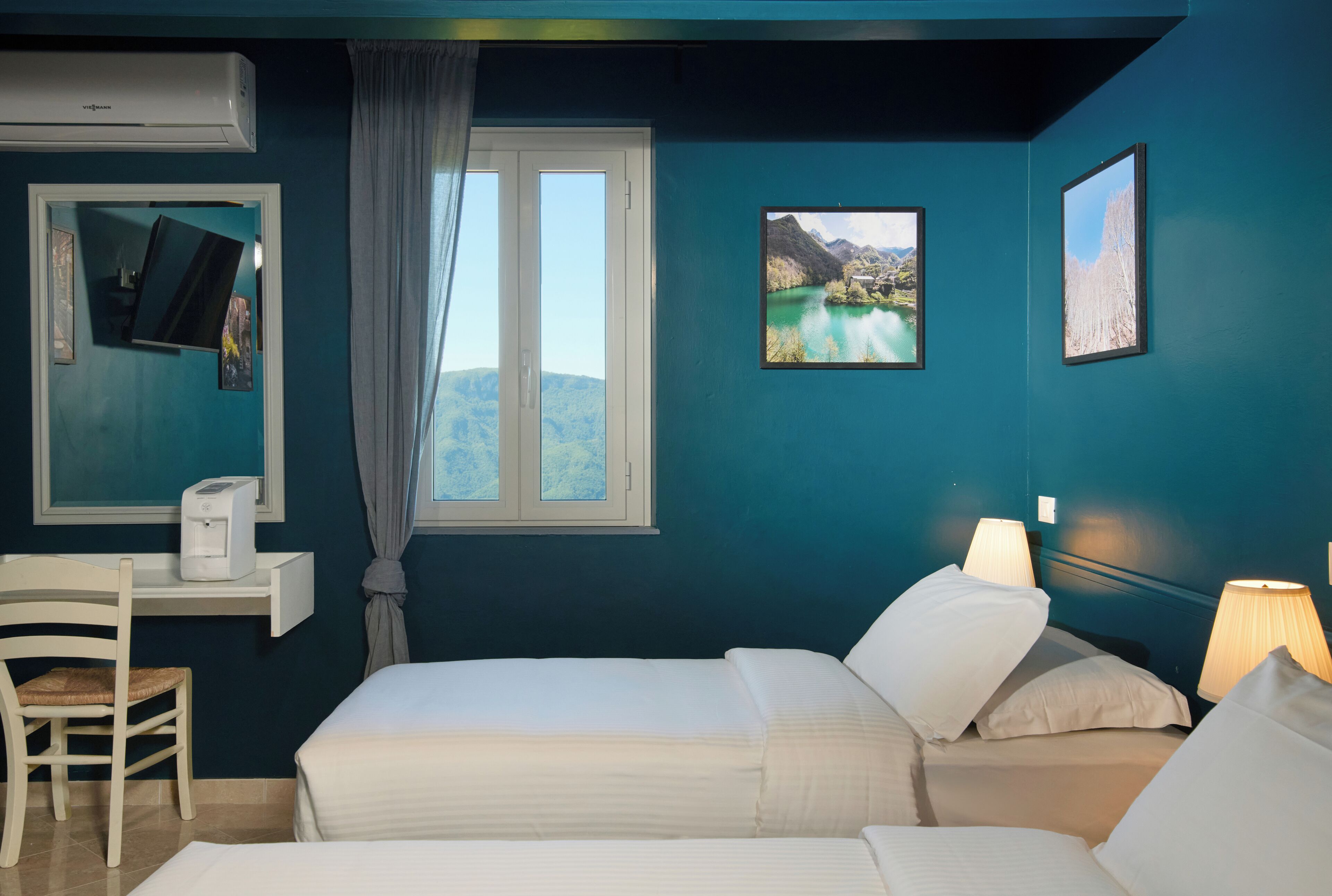 Ein Zimmer mit zwei Betten, einem Schreibtisch mit Computer, einem Stuhl und einem Fenster mit Bergblick.