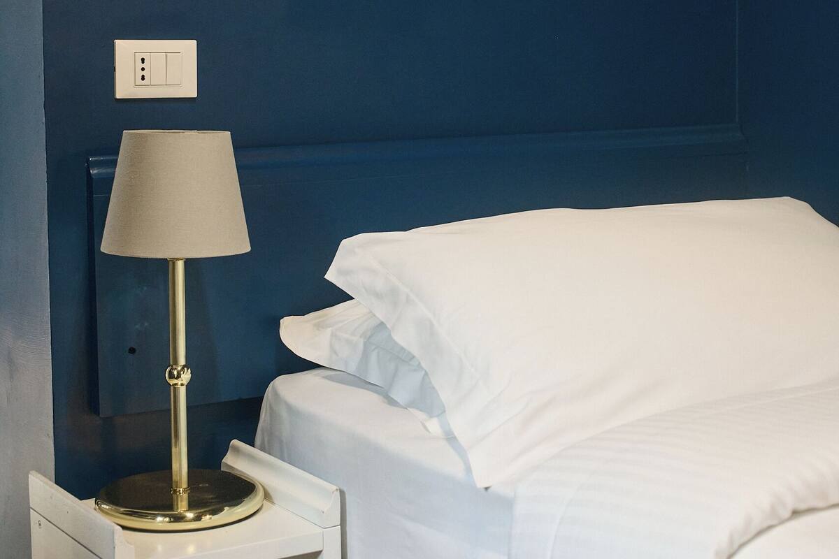 A hotel room with a bed, a nightstand with a lamp, a wall-mounted power outlet, and a framed picture above the bed.