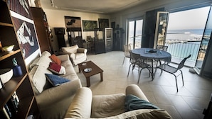 Ein geräumiges Wohnzimmer mit Sofa, Couchtisch und Essbereich. Ein großes Fenster bietet Blick auf das Meer und einen Yachthafen.