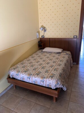 A single bed with a floral-patterned bedspread in a room with a wooden headboard, a nightstand with a lamp, and a wall with a leaf pattern.