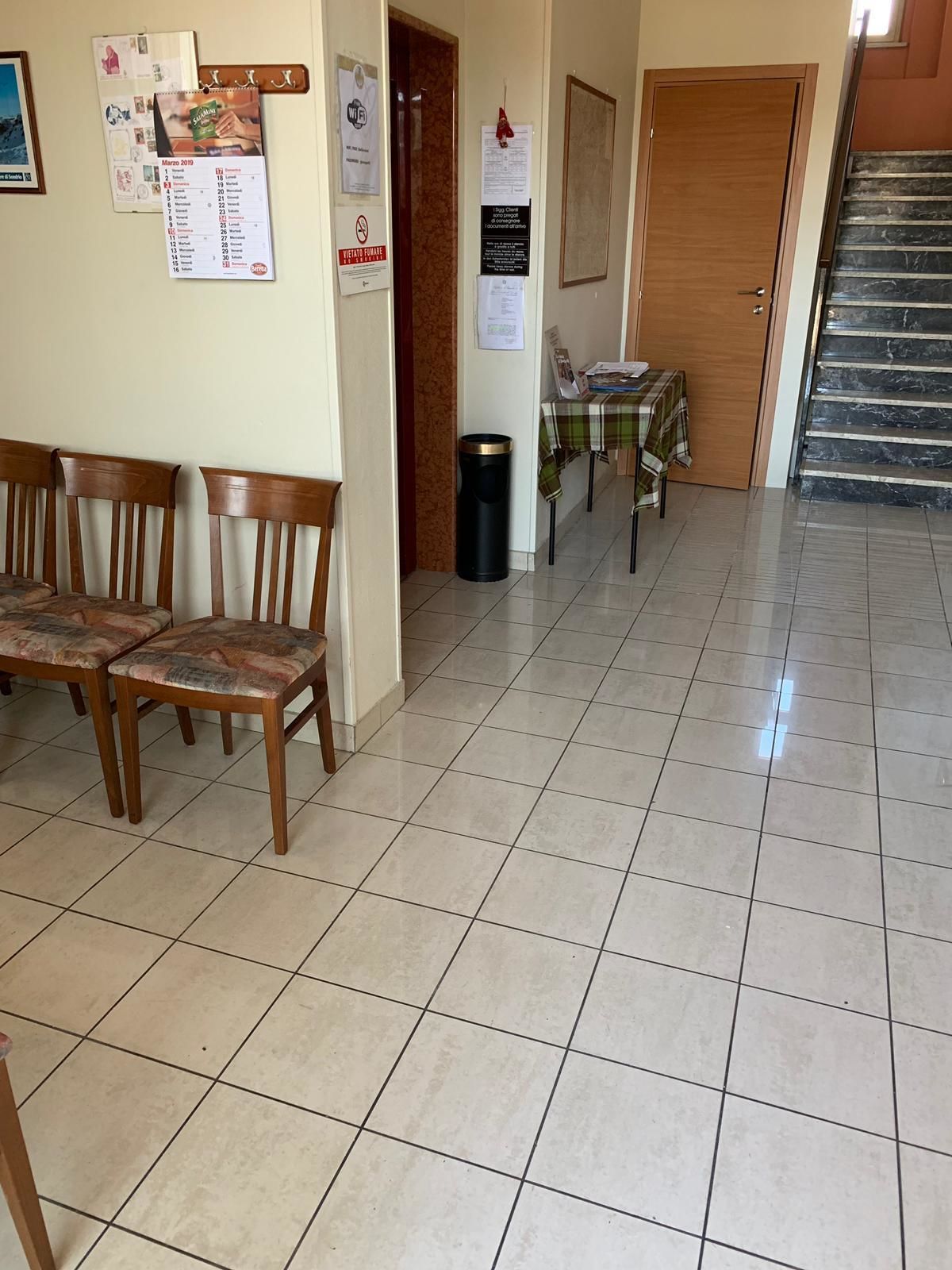 A dining area with wooden chairs and a table, a trash bin, and a calendar on the wall.