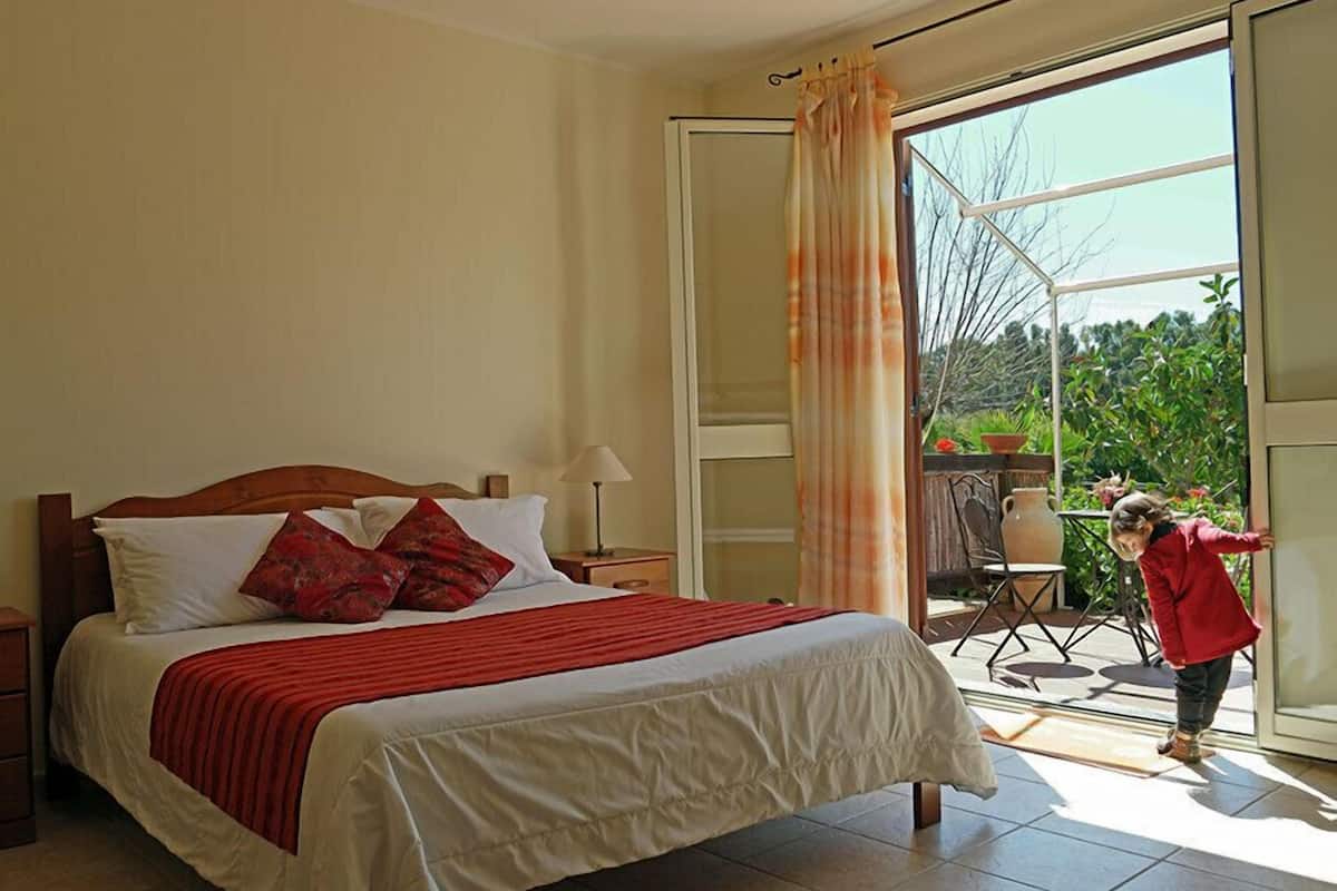 A bedroom with a large bed, a nightstand, a lamp, and a view of a balcony with a person outside.