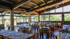 Un restaurante con varias mesas cubiertas con manteles a cuadros, sillas de madera y un ventilador de techo. Amplias ventanas dejan entrar luz natural y ofrecen vistas a zonas verdes exteriores.