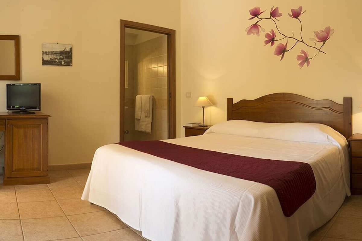 A hotel room with a bed, a television on a wooden cabinet, a bathroom door, and a wall with a floral painting.