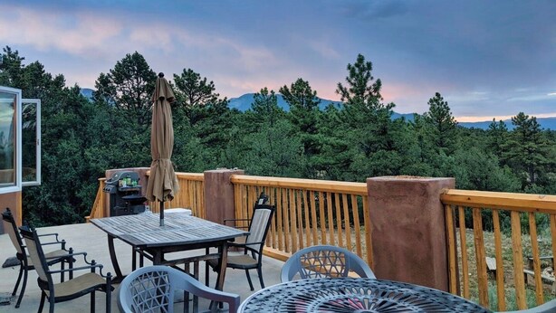 Eine Holzterrasse mit Tisch, Stühlen, Grill und Blick auf Wald und Berge.