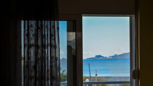 A view from a room with a sliding glass door leading to a balcony overlooking a coastal area with mountains in the distance. - Tommy's Rooms (Rhodes)