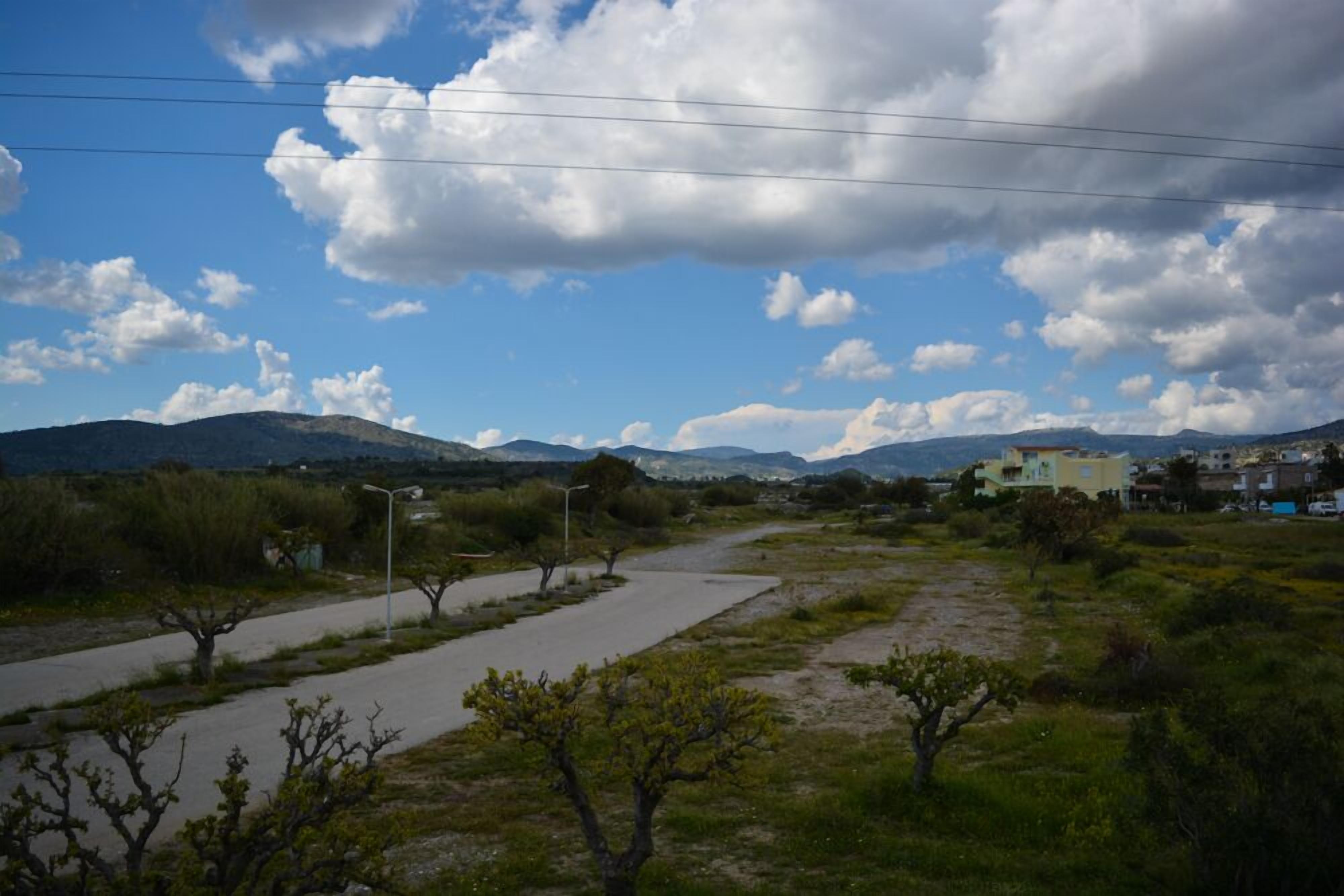 Un paysage rural avec une route asphaltée, une végétation clairsemée et des montagnes lointaines sous un ciel partiellement nuageux.