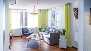 Un salon moderne avec un canapé, un fauteuil et une table basse. De grandes fenêtres sont ornées de rideaux, un lustre illumine la pièce et une horloge est accrochée au mur.