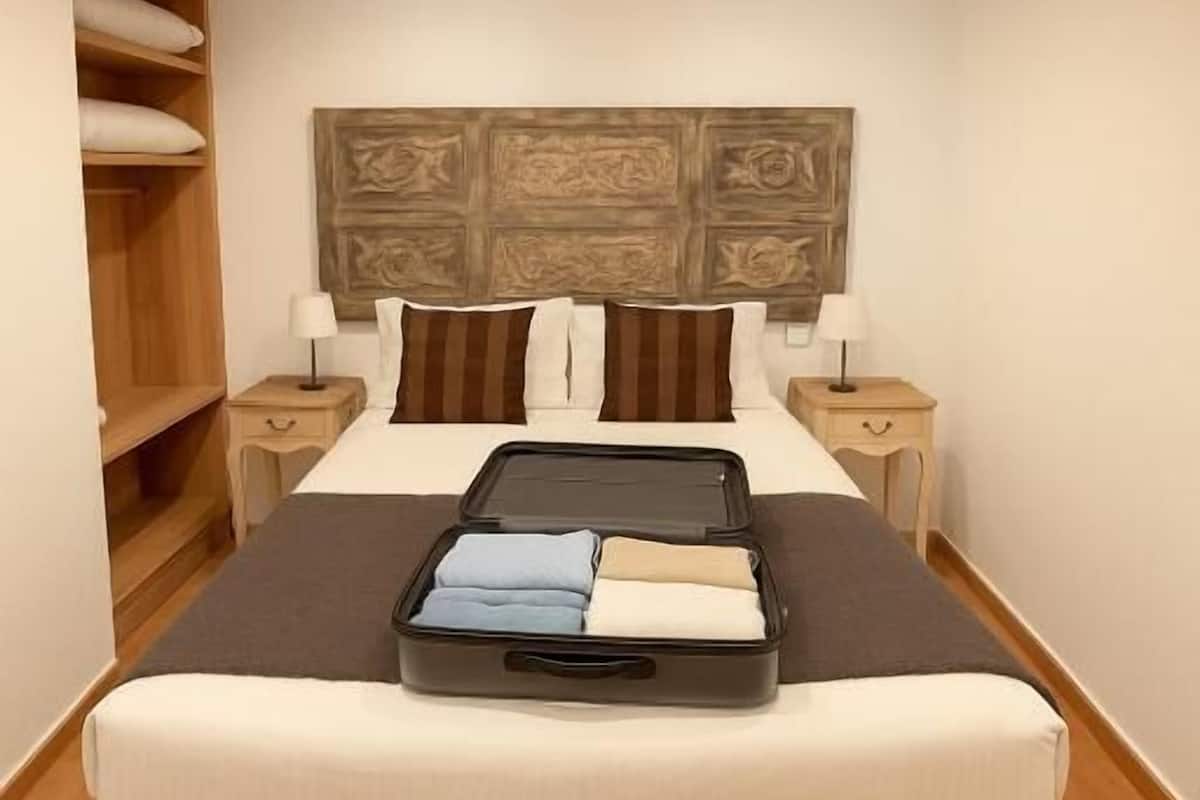 A neatly made bed with a headboard, two bedside tables, a wooden wardrobe, and a suitcase on the floor.