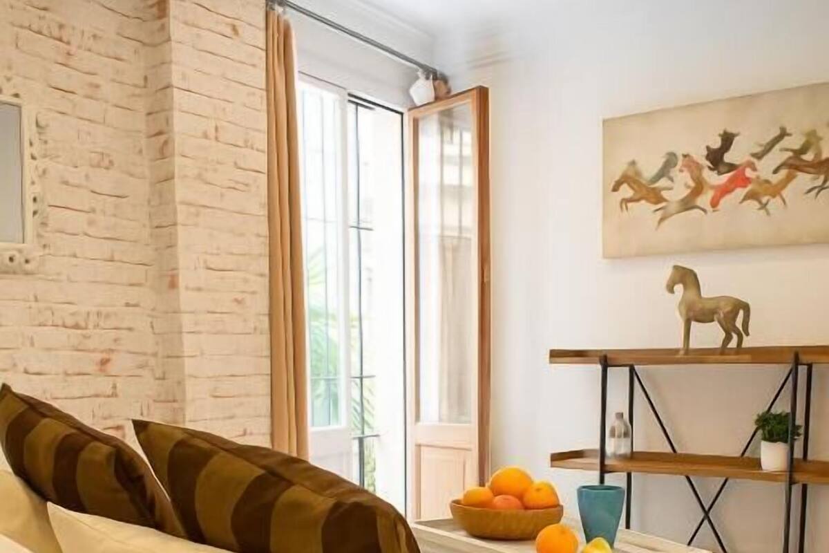 A bedroom with a bed, pillows, a bedside table with a bowl of oranges, a window, and a painting on the wall.
