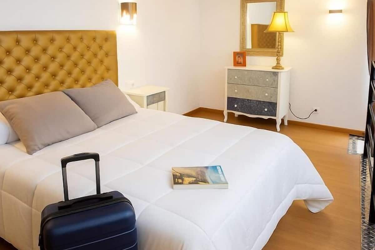 A neatly made bed with a headboard, a bedside table, a lamp, a dresser, and a suitcase on the floor.