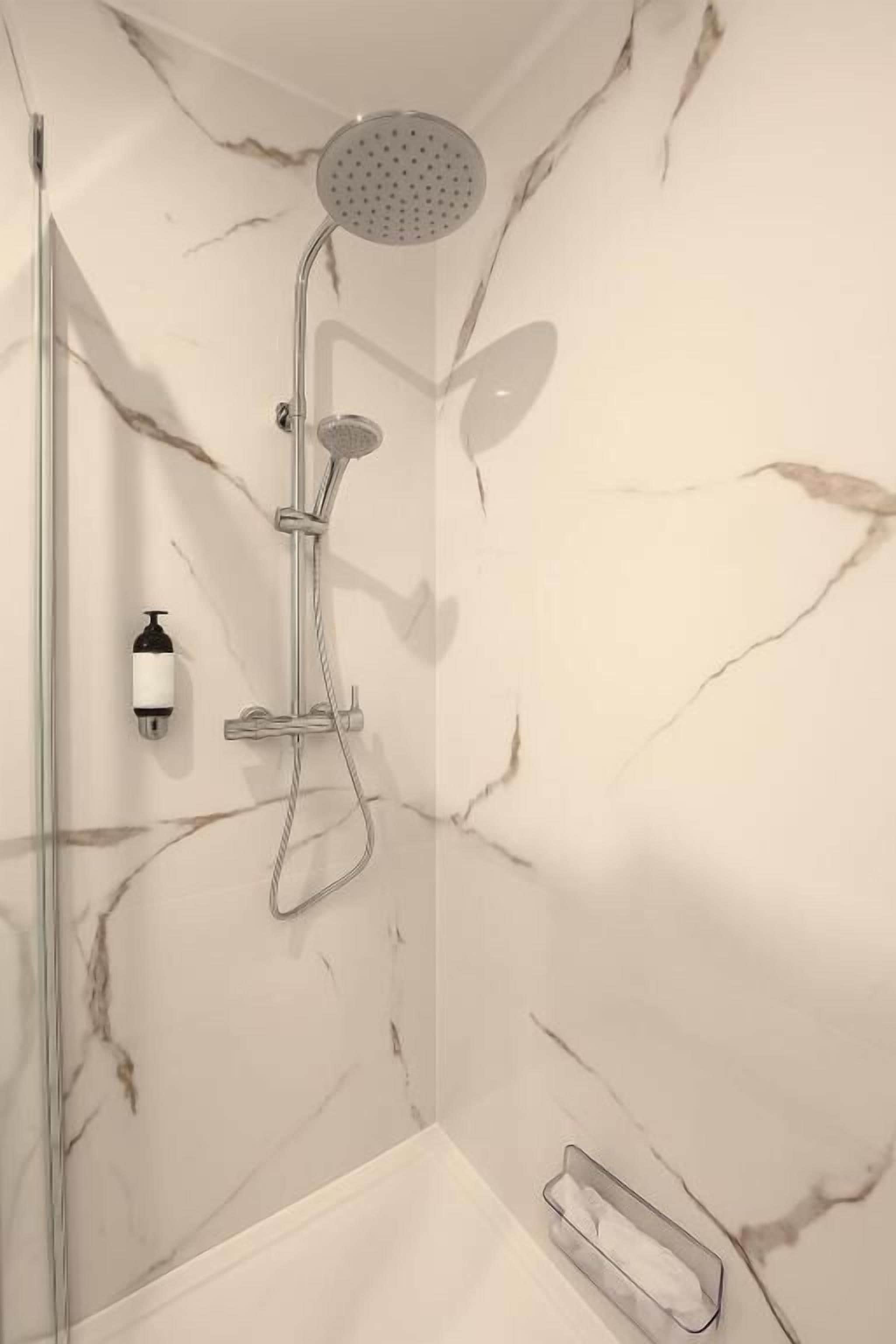 A shower with a handheld showerhead and a wall-mounted rainhead, a soap dispenser, and a glass shower door.