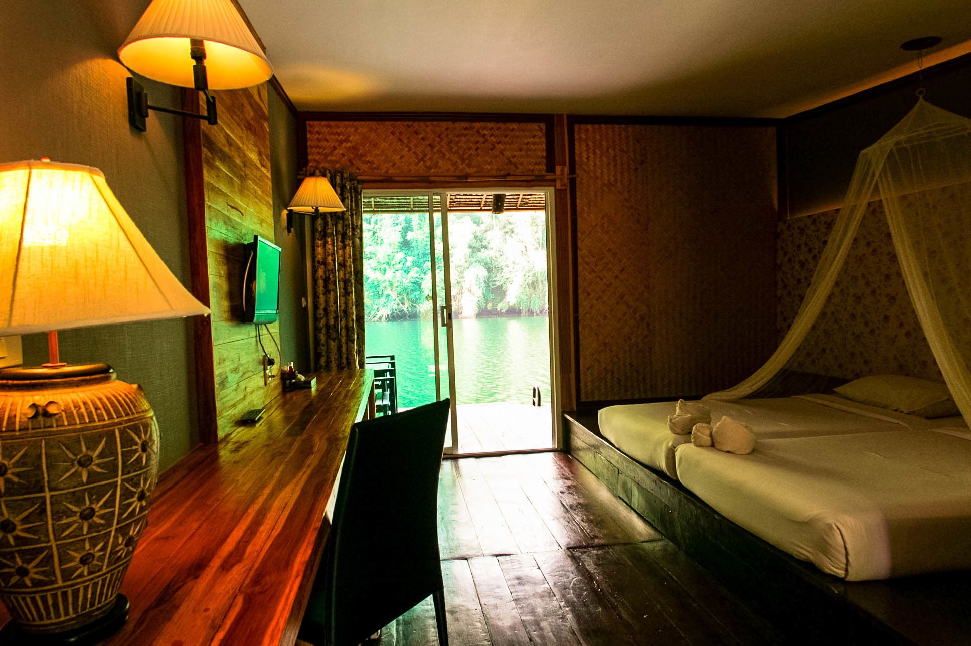 A hotel room with a wooden desk, two lamps, a bed with a canopy, and a view of a body of water through a glass door.