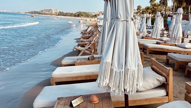 A beachside lounge area with wooden loungers and white umbrellas, a reserved sign, and a view of the ocean and distant buildings. - Kalia Beach Hotel Side  (Manavgat)