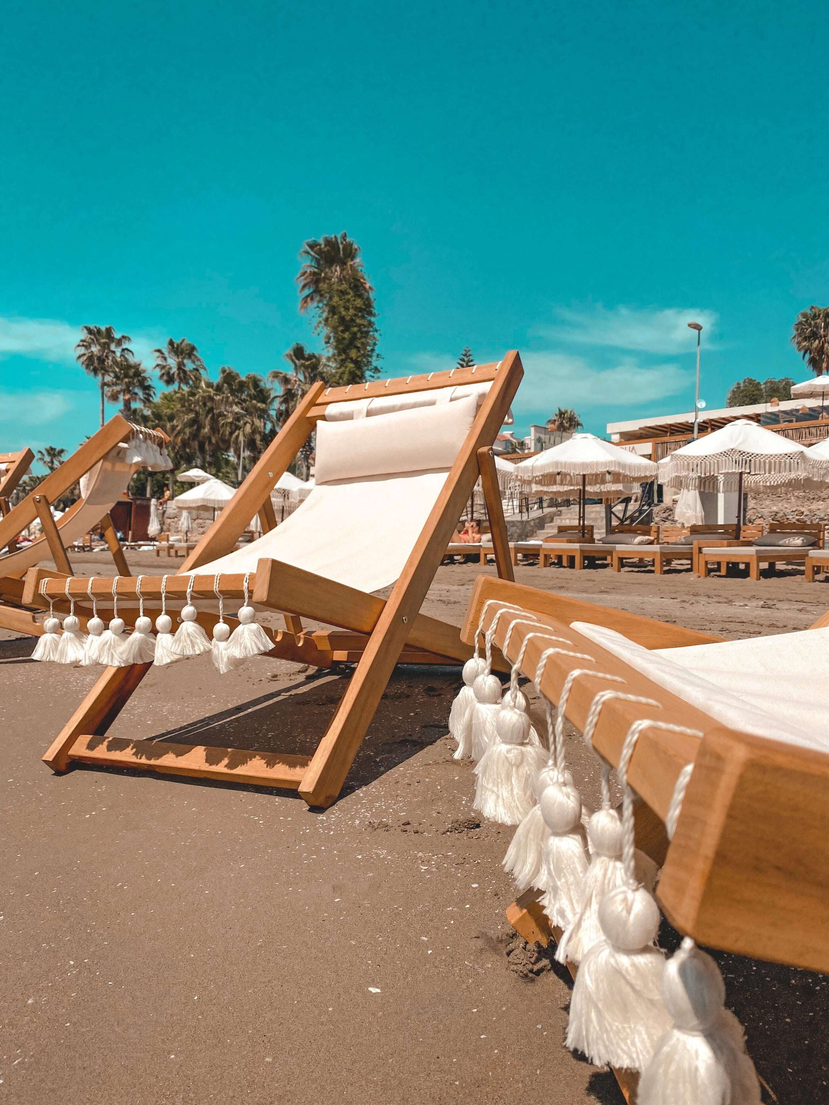Beach chairs with white cushions and tassels, arranged on a sandy beach with palm trees in the background.