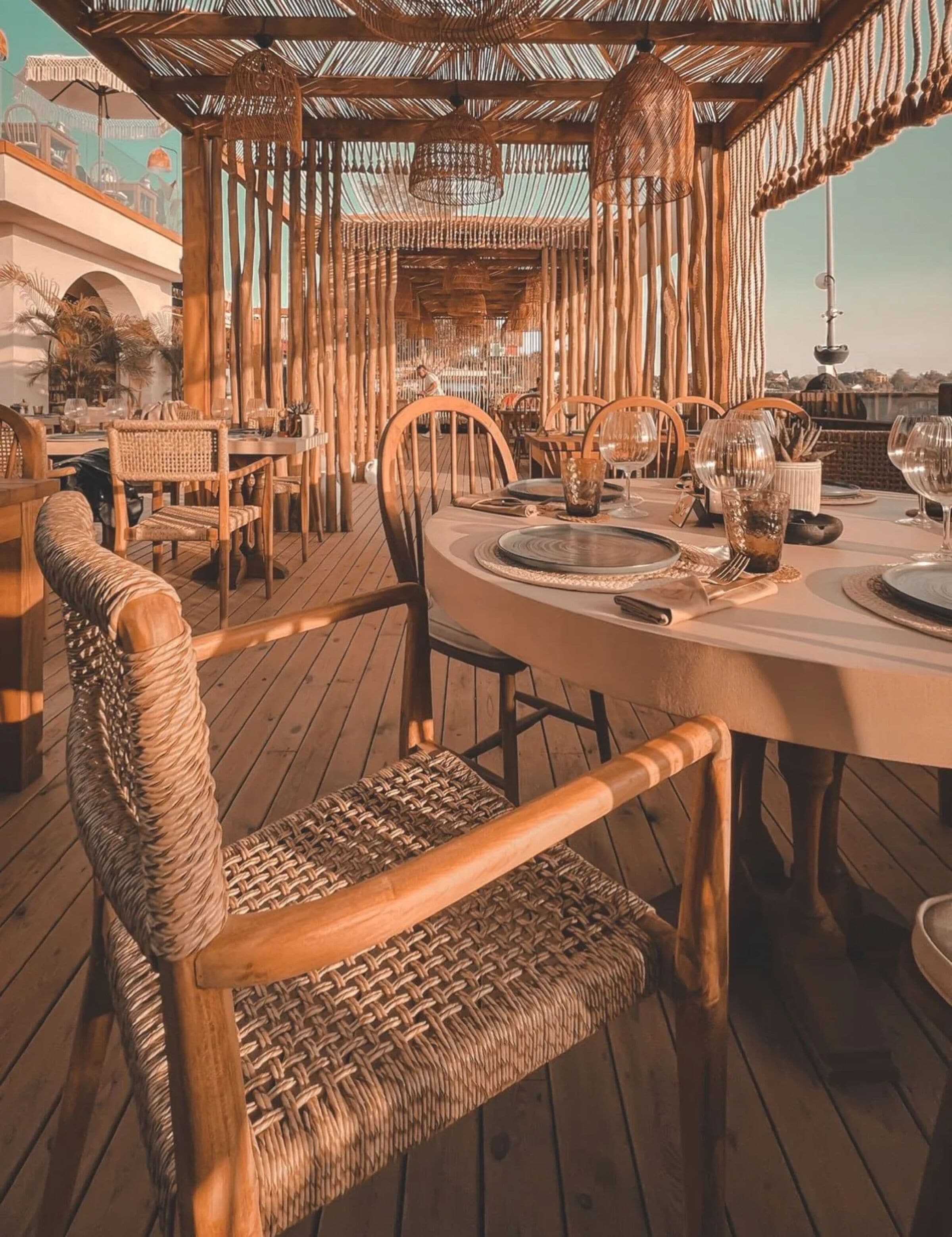 Una terraza de madera con mesas redondas y sillas de mimbre, con un techo de paja.