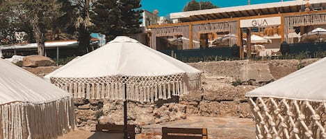 A beach with wooden lounge chairs and white umbrellas, a building with 'GUA' signage, and palm trees in the background.
