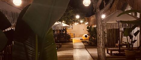 Un sendero de madera conduce a una edificación con techo de paja, rodeada de plantas tropicales e iluminada por luces colgantes.