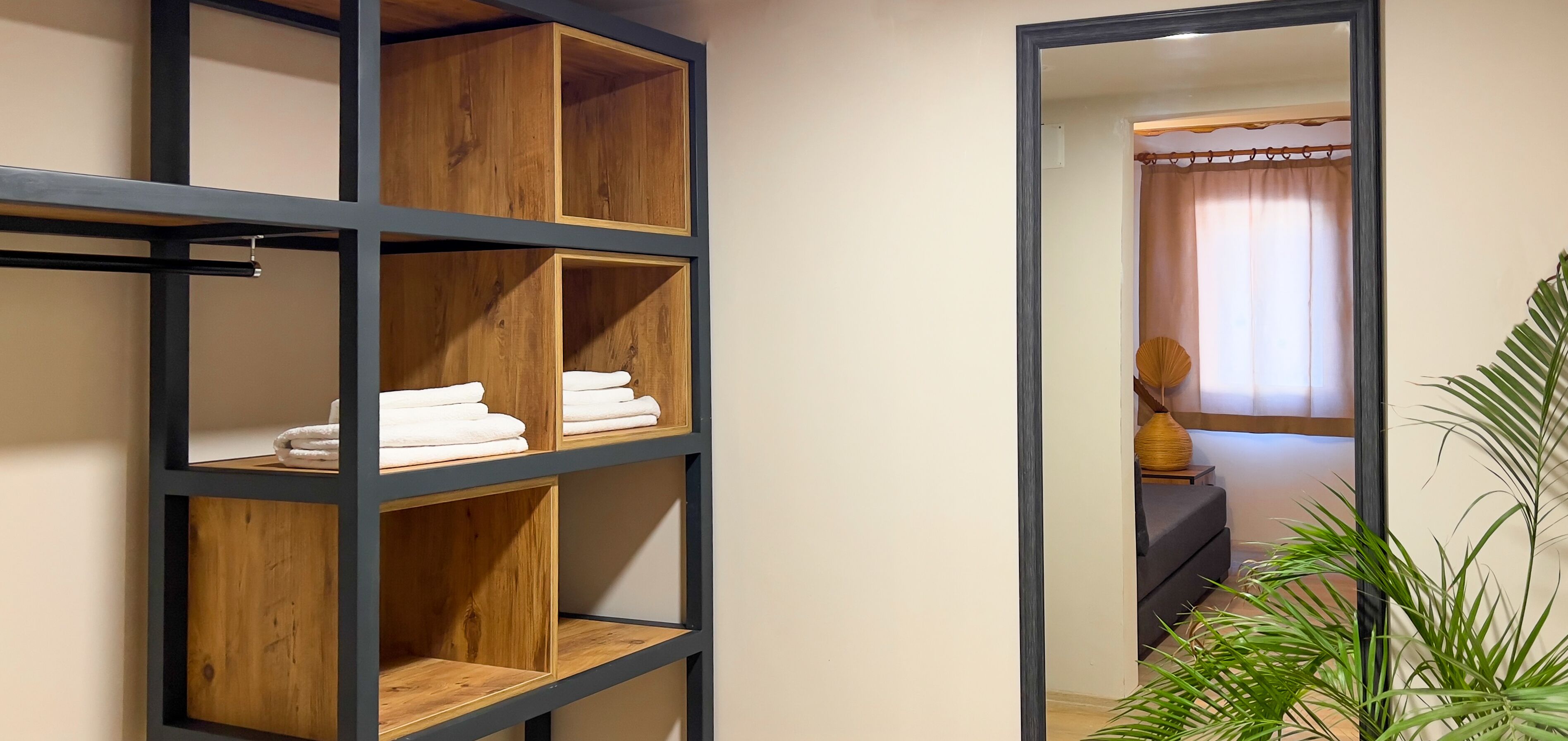 A room with a wooden shelving unit against the wall, a mirror reflecting a bathroom with a shower curtain and a towel rack.