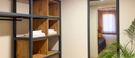 A room with a wooden shelving unit against the wall, a mirror reflecting a bathroom with a shower curtain and a towel rack.