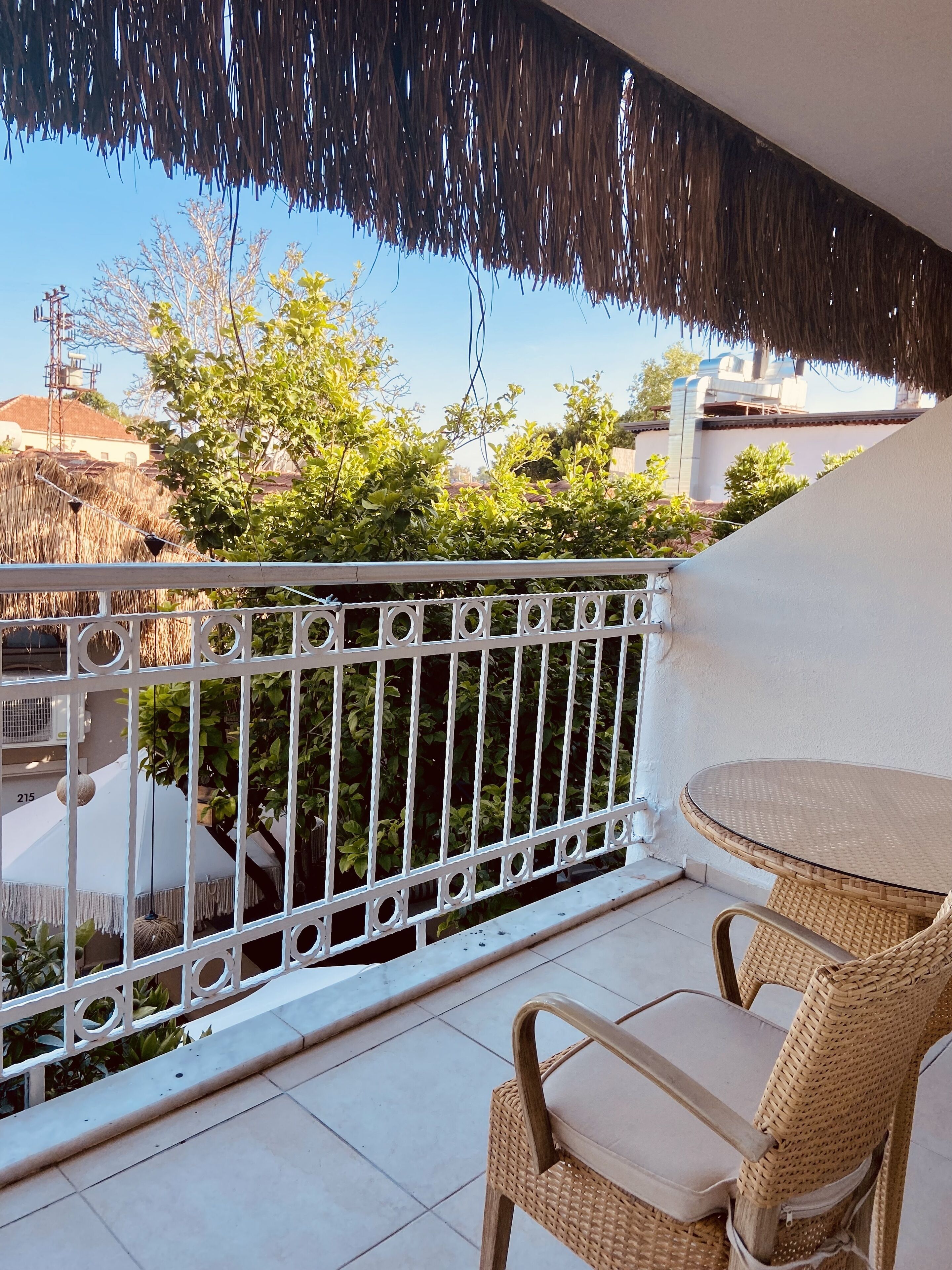 A balcony with a wicker chair and table, white railing, and a view of greenery and buildings.