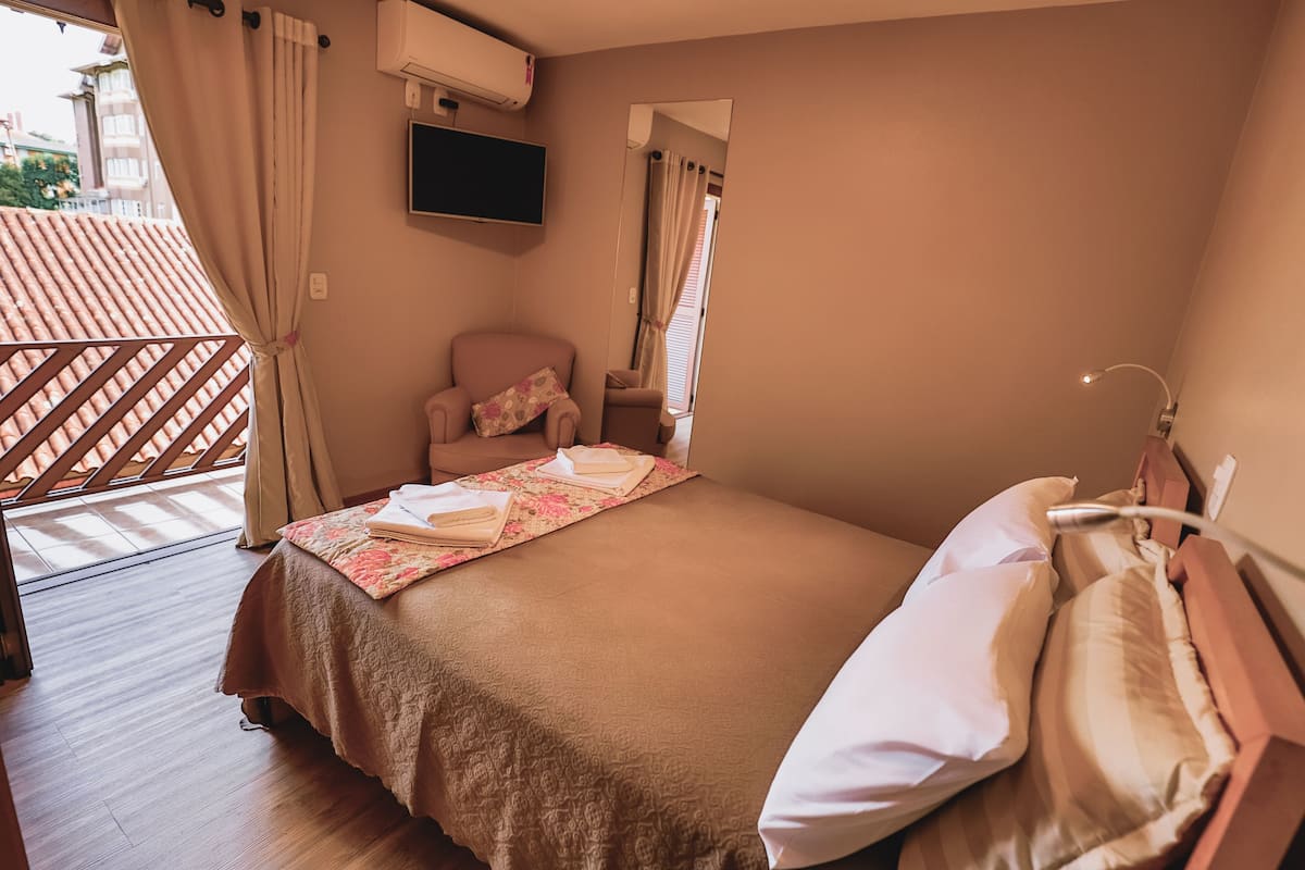 Ein ordentlich bezogenes Bett mit Blumenmuster, ein Sessel mit Kissen und ein an der Wand montierter Fernseher.