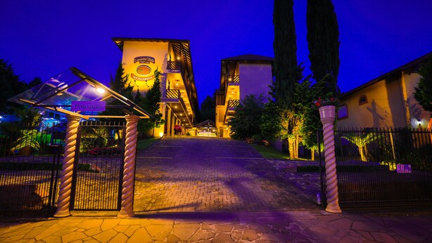 Entrada de hotel con un camino pavimentado que conduce a una puerta con portones, rodeada de árboles altos y una fachada de edificio bien iluminada.