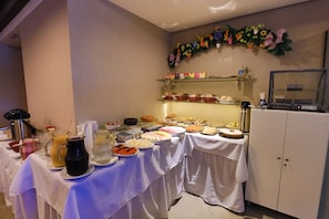 Una mesa de buffet con diversos alimentos y bebidas, una pared decorada con flores, y un mueble con puerta de vidrio.