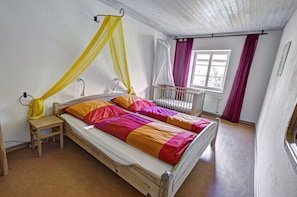 A single bed with a canopy, a small wooden stool, a crib, and a window with curtains.
