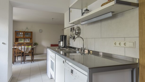A modern kitchen with a stainless steel island, a built-in oven, and a coffee maker. There is a dining area with wooden chairs and a small table. - Nengshof (Wissmannsdorf)