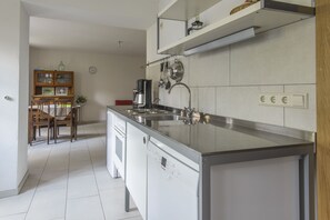 A modern kitchen with a stainless steel island, a built-in oven, and a coffee maker. There is a dining area with wooden chairs and a small table. - Nengshof (Wissmannsdorf)