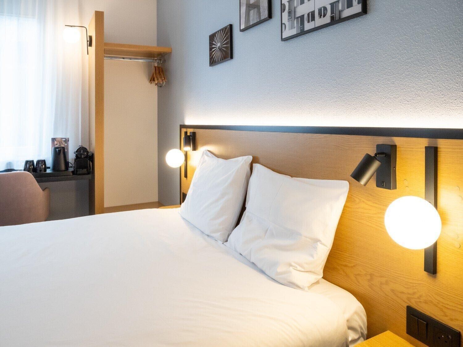 Habitación de hotel con una cama grande, lámparas de noche, cabecera de madera y un pequeño escritorio con cafetera.