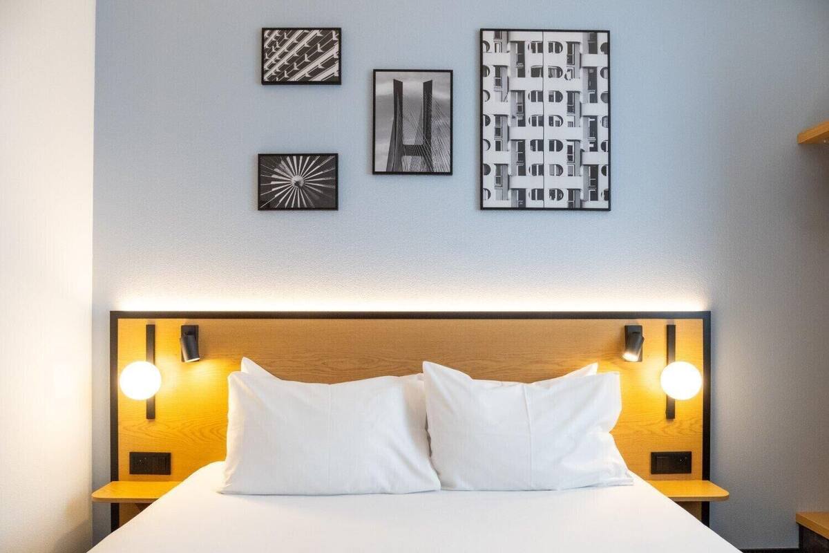 A neatly made bed with two pillows, a wooden headboard, and wall-mounted lights.