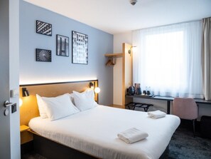 A hotel room with a large bed, a desk, and a chair. There are framed pictures on the wall and a window with curtains.