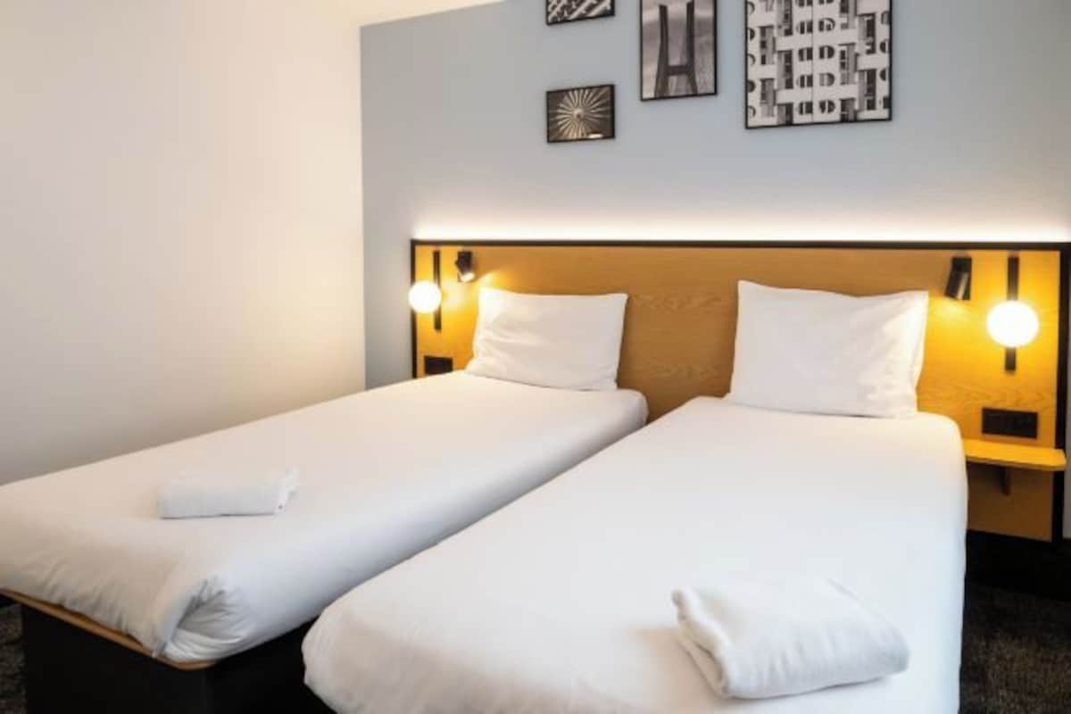 A hotel room with two single beds, each with a white duvet and pillow, and a headboard with integrated wall sconces.