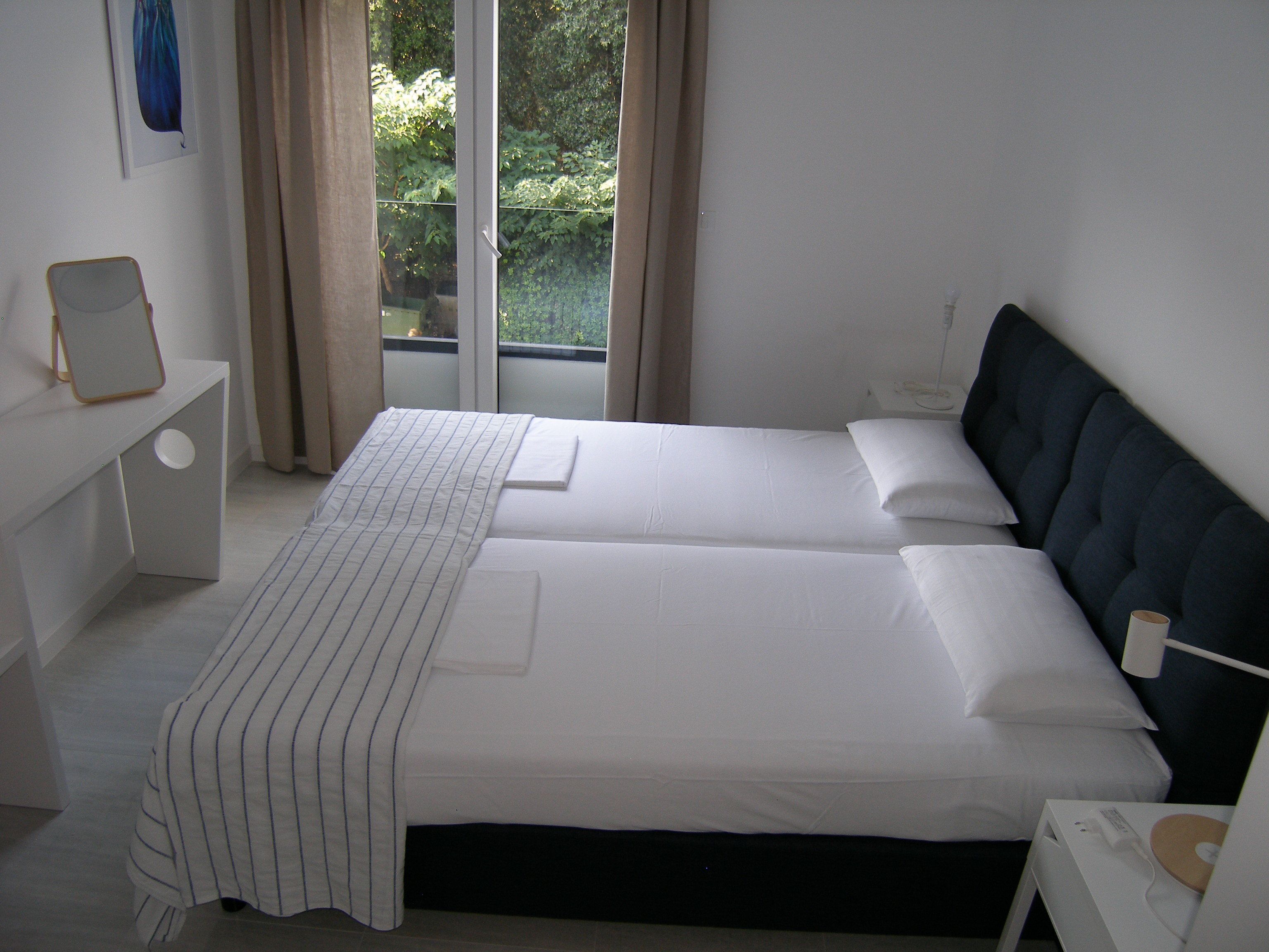 A double bed with white linens, a bedside table with a lamp, and a view of greenery through a window.