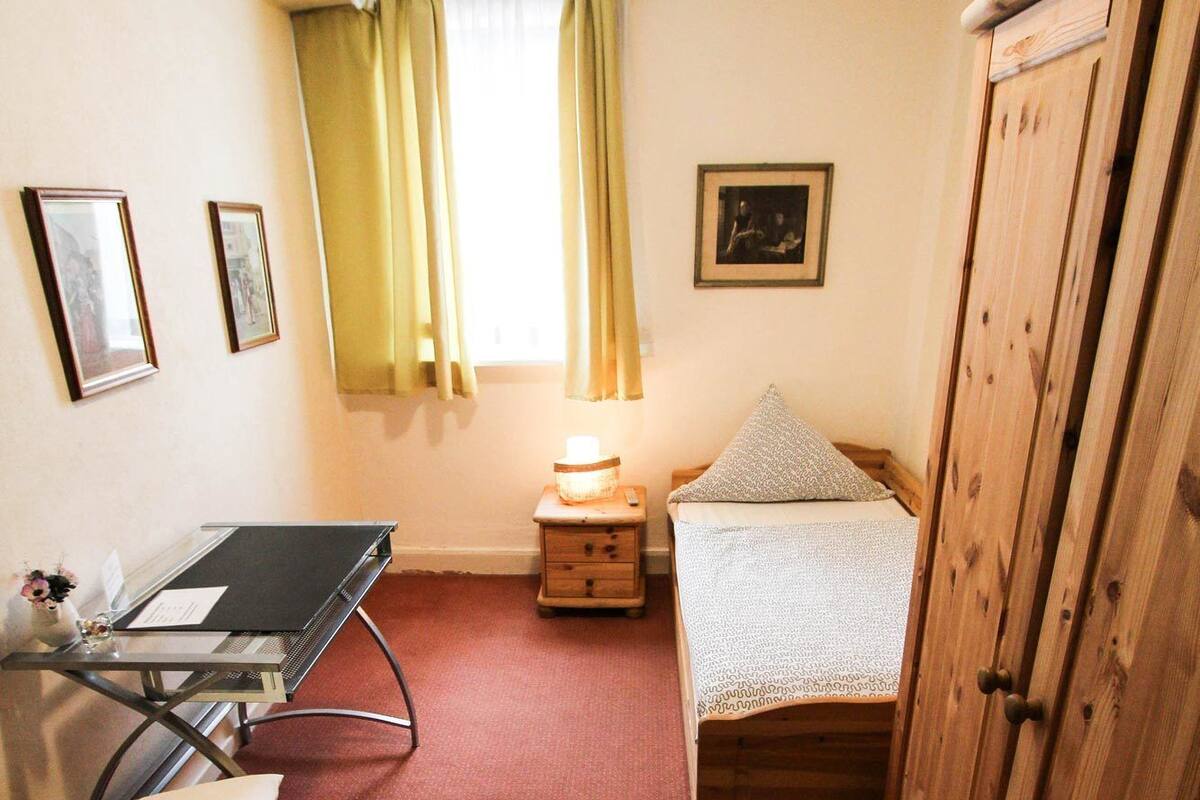 Een kleine kamer met een bed, een bureau met stoel, een kledingkast en een klein nachtkastje met lamp.