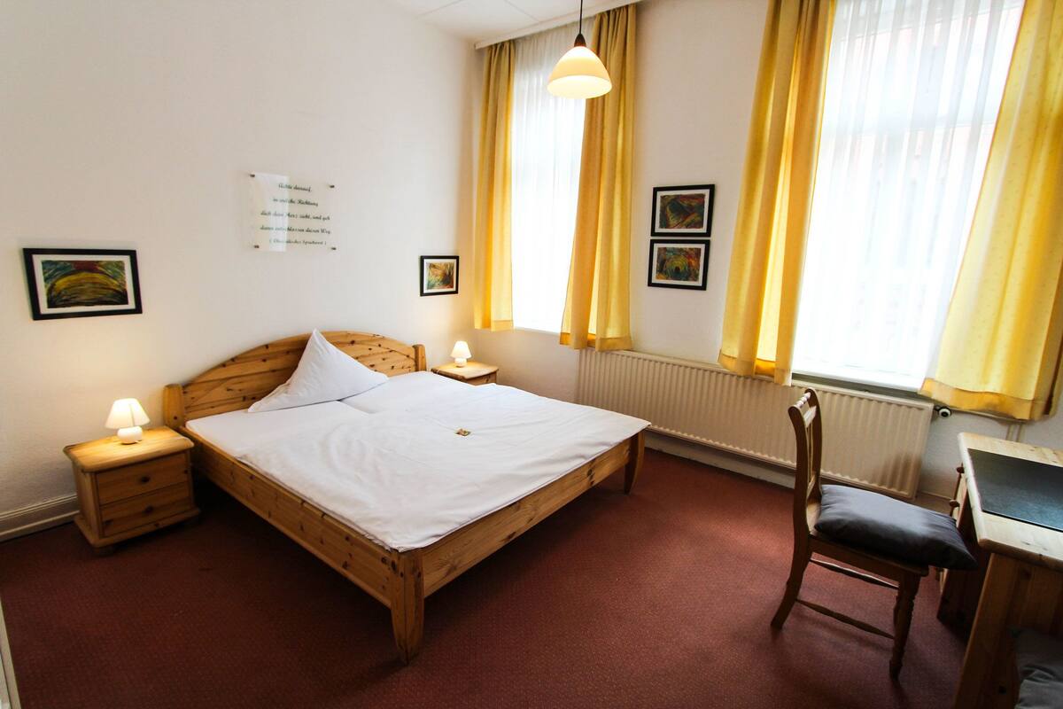 Een kamer met een houten bed, een stoel, een bureau en een raam met gordijnen.