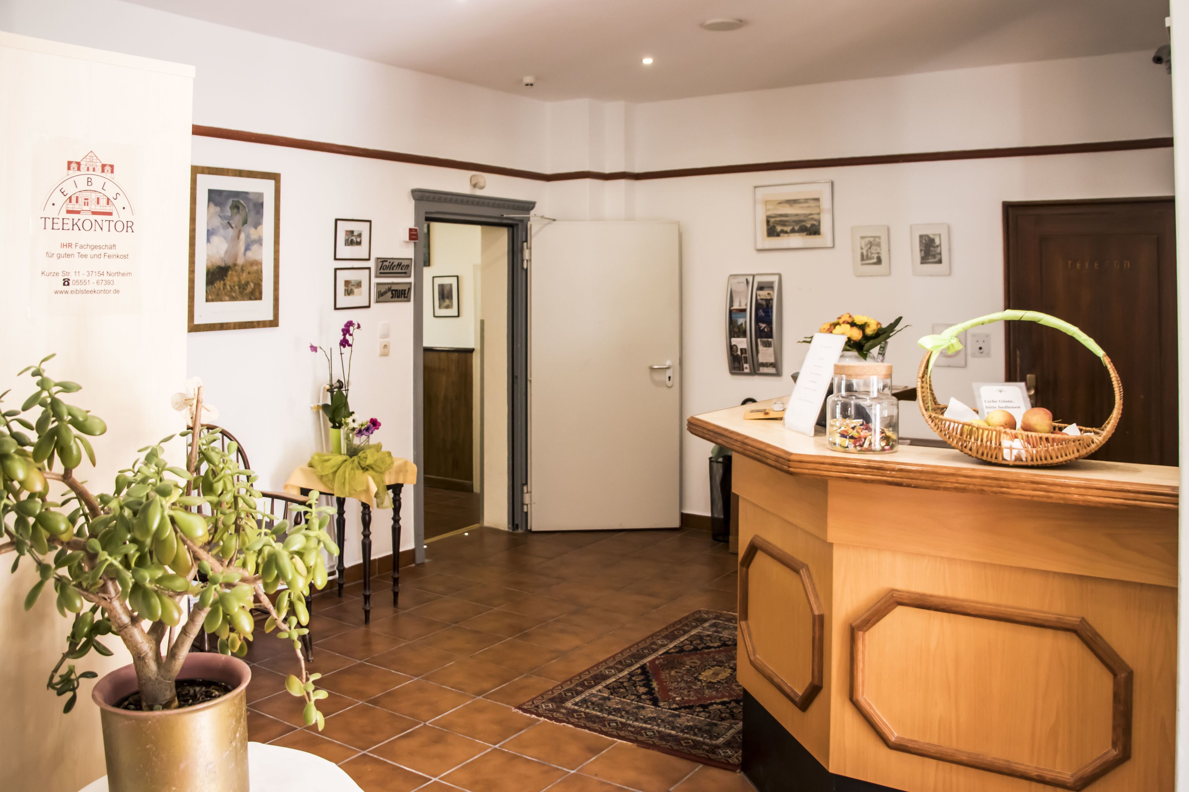 A reception area with a wooden reception desk, a potted plant, framed pictures on the wall, and a tiled floor.