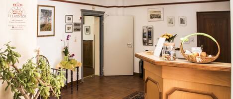 A reception area with a wooden reception desk, a potted plant, framed pictures on the wall, and a tiled floor.