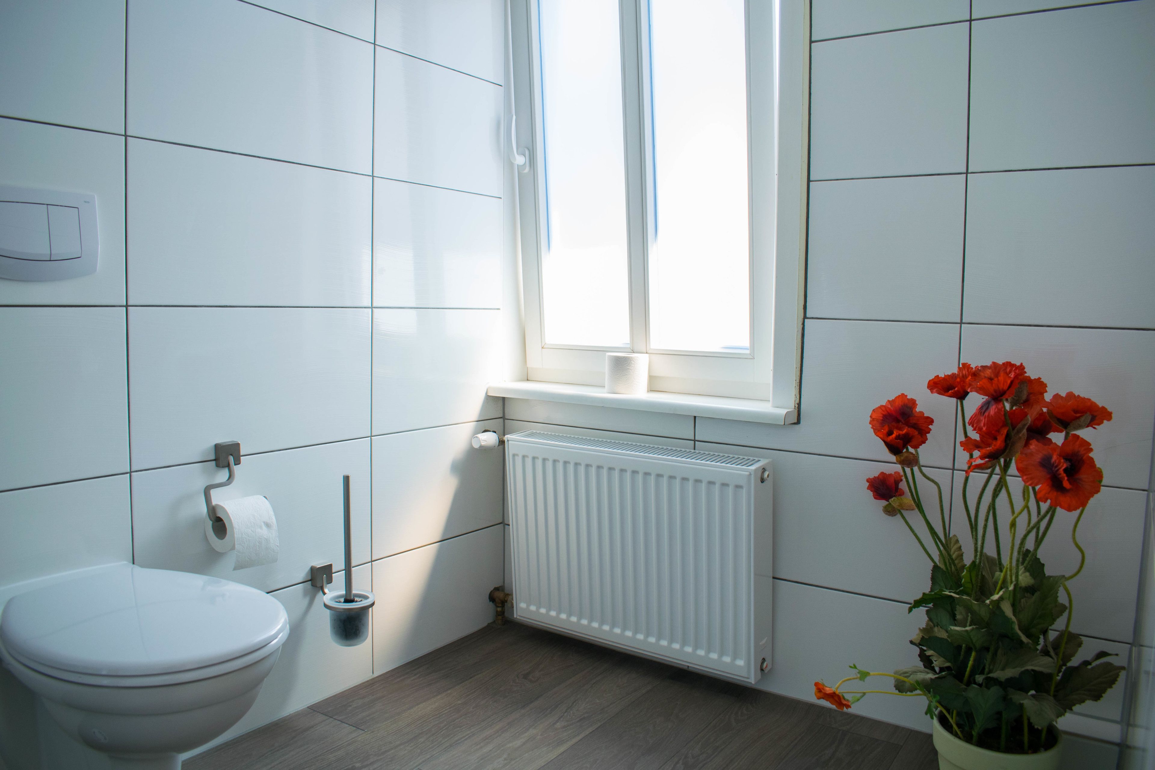 A bathroom with a toilet, a radiator, a window, and a potted plant with flowers.