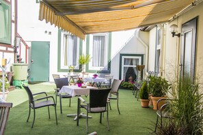 Une terrasse extérieure avec des tables et des chaises, entourée de plantes en pot et d’un auvent à rayures.
